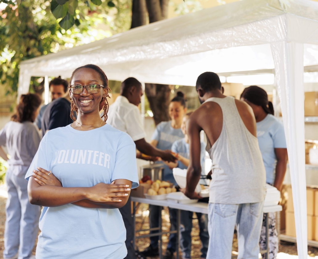 Young black woman with glasses stands outdoors, arms crossed, looking at the camera. Diverse group of volunteers supports a non-profit program dedicated to hunger relief and helping needy individuals.